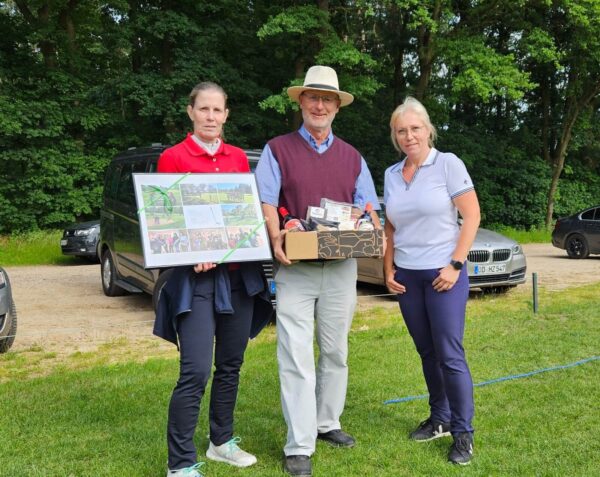 Abschied Sven Busch Jugendtrainer Drei Personen stehen auf einer Wiese vor einem Wald. Eine Person in Rot hält einen Bilderrahmen, die mittlere Person mit Hut und Weste hält eine Geschenkbox, und die dritte Person in Weiß steht daneben. Im Hintergrund sind Autos geparkt.