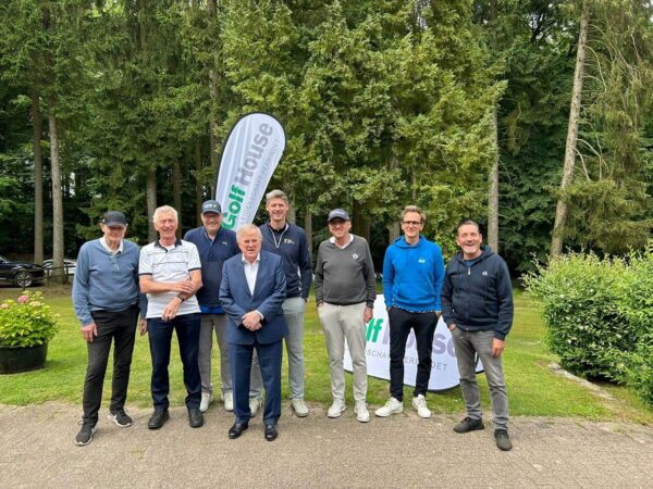 Ehrengäste 6.ProAm GC Hoisdorf Eine Gruppe von acht Personen steht lächelnd für ein Foto auf einem Weg vor einer bewaldeten Kulisse. Hinter ihnen ist ein Werbebanner zu sehen. Einige tragen sportliche Kleidung, andere Anzüge.