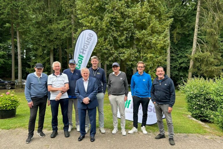 Eine Gruppe von acht Personen steht lächelnd für ein Foto auf einem Weg vor einer bewaldeten Kulisse. Hinter ihnen ist ein Werbebanner zu sehen. Einige tragen sportliche Kleidung, andere Anzüge.