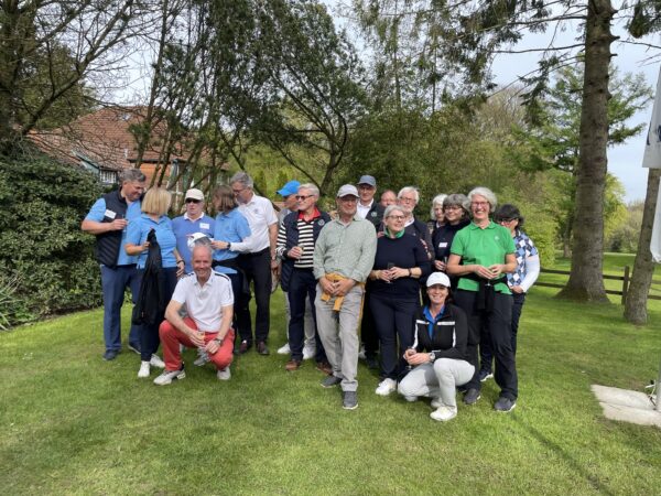 Helferteam Golferlebnistag Eine Gruppe von Menschen posiert lächelnd im Freien auf einer grünen Wiese vor Bäumen. Einige tragen Freizeitkleidung, andere Golfkleidung. Im Hintergrund sind Büsche und ein Gebäude zu sehen.
