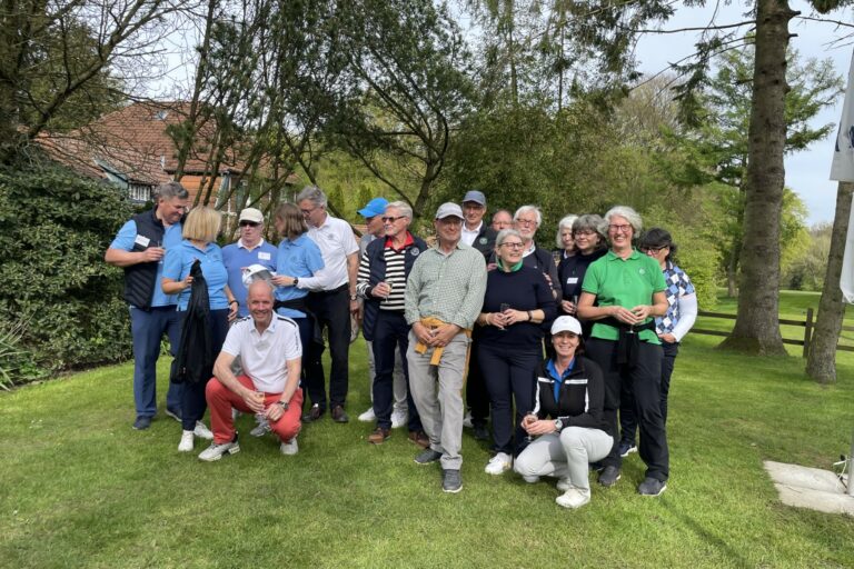 Eine Gruppe von Menschen posiert lächelnd im Freien auf einer grünen Wiese vor Bäumen. Einige tragen Freizeitkleidung, andere Golfkleidung. Im Hintergrund sind Büsche und ein Gebäude zu sehen.
