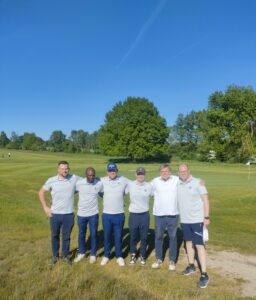 Herrren Mannschaft Neu Sechs Personen stehen auf einem Golfplatz nebeneinander und lächeln in die Kamera. Alle tragen Poloshirts und dunkle Hosen. Im Hintergrund befinden sich Bäume und ein blauer Himmel.