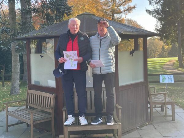 Zwei ältere Männer stehen auf einer Bank vor einem kleinen Holzgebäude im Freien. Sie tragen Jacken und halten Papierblätter in den Händen. Im Hintergrund sind Bäume und ein Golfplatz zu sehen. Einer der Männer hebt die Hand an die Stirn.