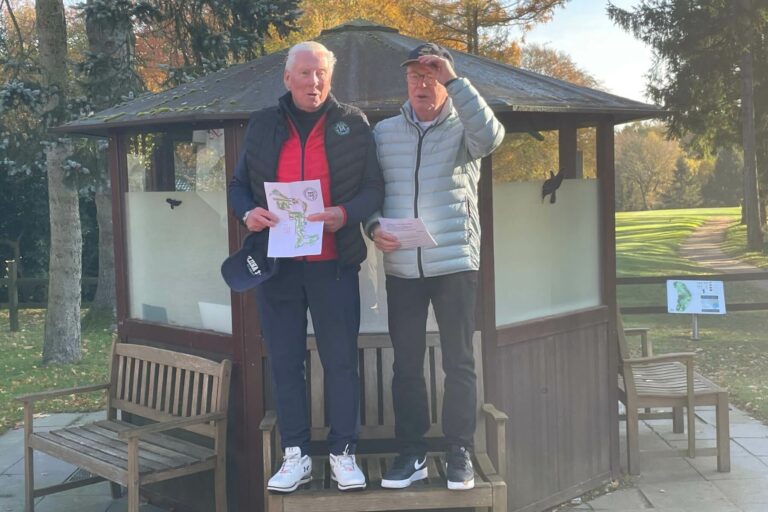 Zwei ältere Männer stehen auf einer Bank vor einem kleinen Holzgebäude im Freien. Sie tragen Jacken und halten Papierblätter in den Händen. Im Hintergrund sind Bäume und ein Golfplatz zu sehen. Einer der Männer hebt die Hand an die Stirn.