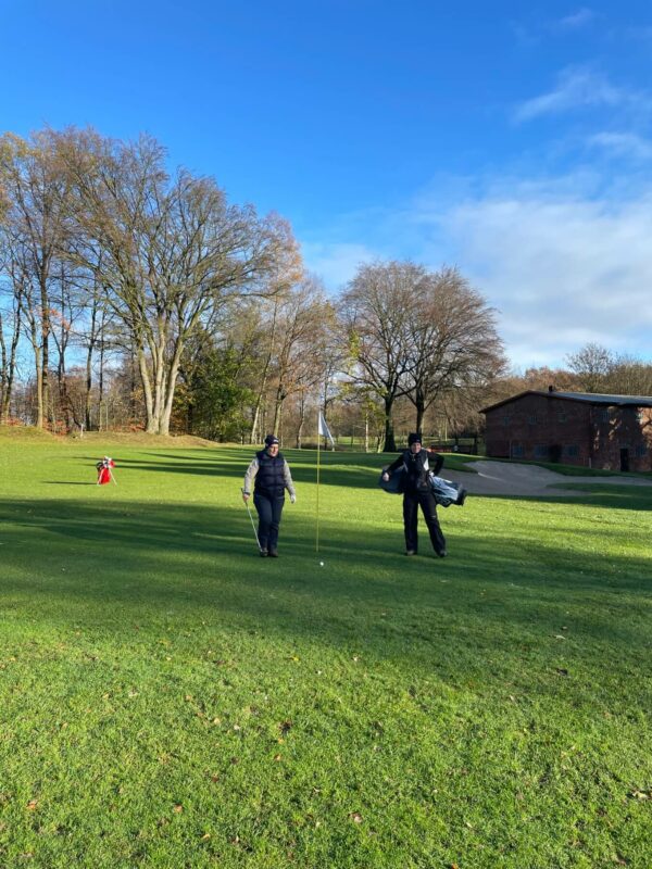 Platz Herbst Golf Club Hoisdorf 2915 Zwei Personen stehen auf einem Golfplatz neben der Fahne eines Lochs. Sie halten Golfschläger und eine Golftasche. Im Hintergrund sind Grasflächen, Bäume ohne Laub und ein Backsteingebäude zu sehen. Der Himmel ist blau mit wenigen Wolken.