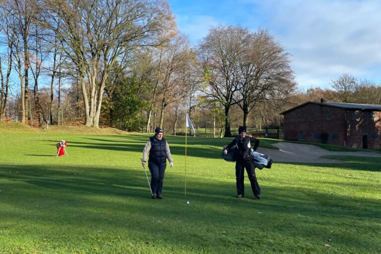 Zwei Personen stehen auf einem Golfplatz neben der Fahne eines Lochs. Sie halten Golfschläger und eine Golftasche. Im Hintergrund sind Grasflächen, Bäume ohne Laub und ein Backsteingebäude zu sehen. Der Himmel ist blau mit wenigen Wolken.