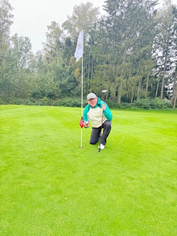 Roland Jahn Hole I N1 25.9 1 Ein Mensch kniet auf einem Golfplatz neben einem Loch mit Fahne. Sie halten einen Golfschläger und einen Golfball, umgeben von grünem Rasen und Bäumen im Hintergrund.