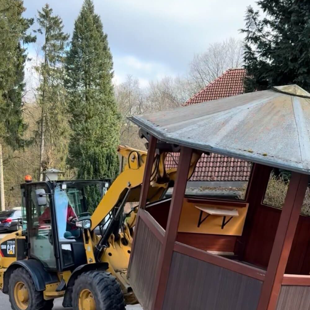 Ein gelber Bagger hebt einen hölzernen Pavillon an. Im Hintergrund sind Bäume und ein Teil eines Daches zu sehen. Der Himmel ist bewölkt.