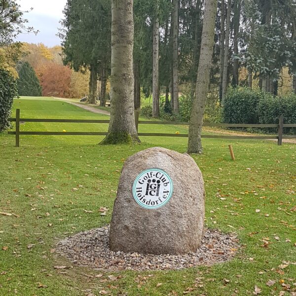 Stein Bahn1 Ein großer Stein mit einem runden Emblem steht auf einer Rasenfläche vor Bäumen. Im Hintergrund sind ein Weg und ein Holzzaun zu sehen. Das Emblem zeigt einen Golfschläger und einen Ball.