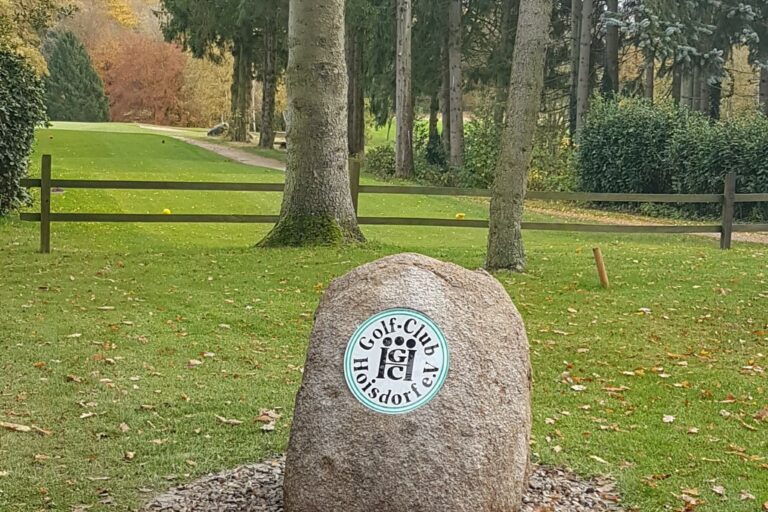 Ein großer Stein mit einem runden Emblem steht auf einer Rasenfläche vor Bäumen. Im Hintergrund sind ein Weg und ein Holzzaun zu sehen. Das Emblem zeigt einen Golfschläger und einen Ball.