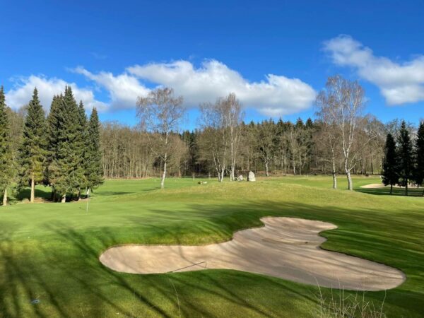 Greenfee 1500w Eine ruhige Golflandschaft unter einem blauen Himmel mit weißen Wolken. Im Vordergrund ein Sandbunker und gepflegter Rasen, umgeben von hohen Nadelbäumen und kahlen Laubbäumen am Horizont.
