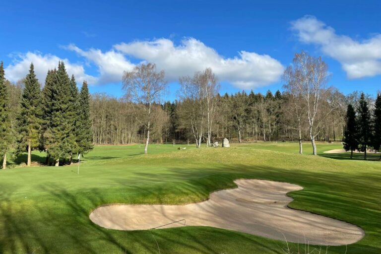 Eine ruhige Golflandschaft unter einem blauen Himmel mit weißen Wolken. Im Vordergrund ein Sandbunker und gepflegter Rasen, umgeben von hohen Nadelbäumen und kahlen Laubbäumen am Horizont.
